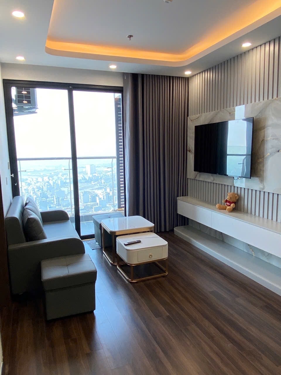 A modern living area features a sleek sofa alongside two minimalist coffee tables. A mounted television is positioned on a stylish wall unit. Floor-to-ceiling windows offer expansive views of the city skyline, with soft curtains framing the space.