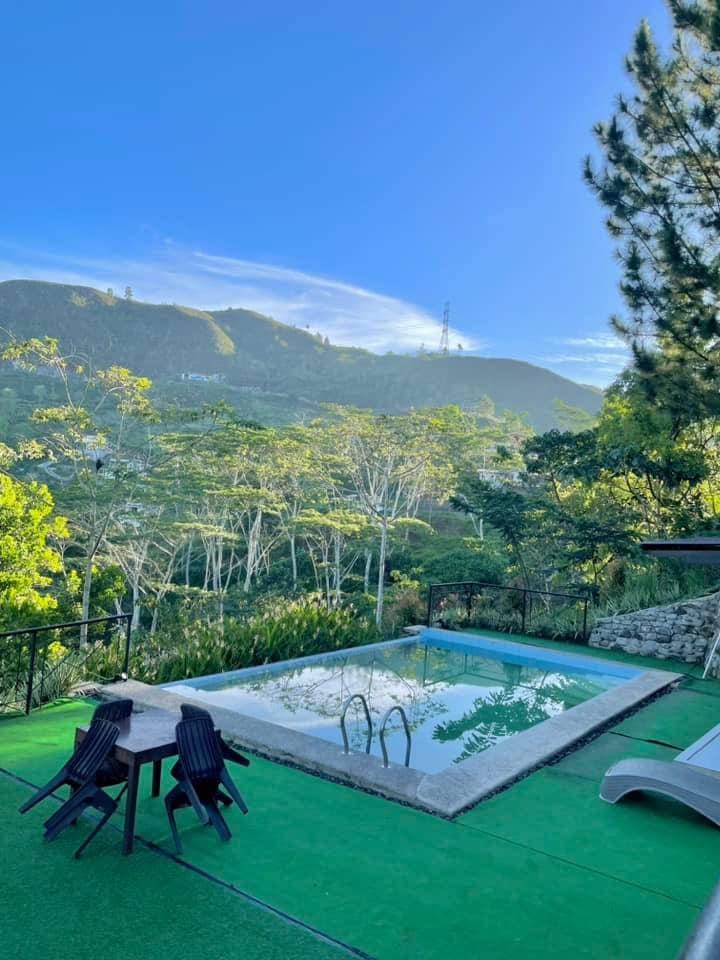 A serene outdoor area is displayed, featuring a tranquil pool surrounded by lush greenery. Two chairs are positioned nearby, offering a perfect spot to enjoy the scenic mountain views under a clear blue sky.