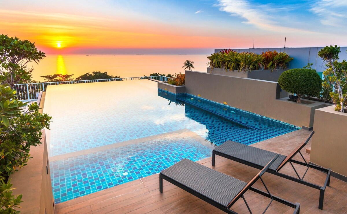 An infinity pool extends towards a sunset, offering panoramic views over the landscape. Stylish lounge chairs are positioned near the water, surrounded by lush greenery and decorative shrubs, enhancing the serene atmosphere.