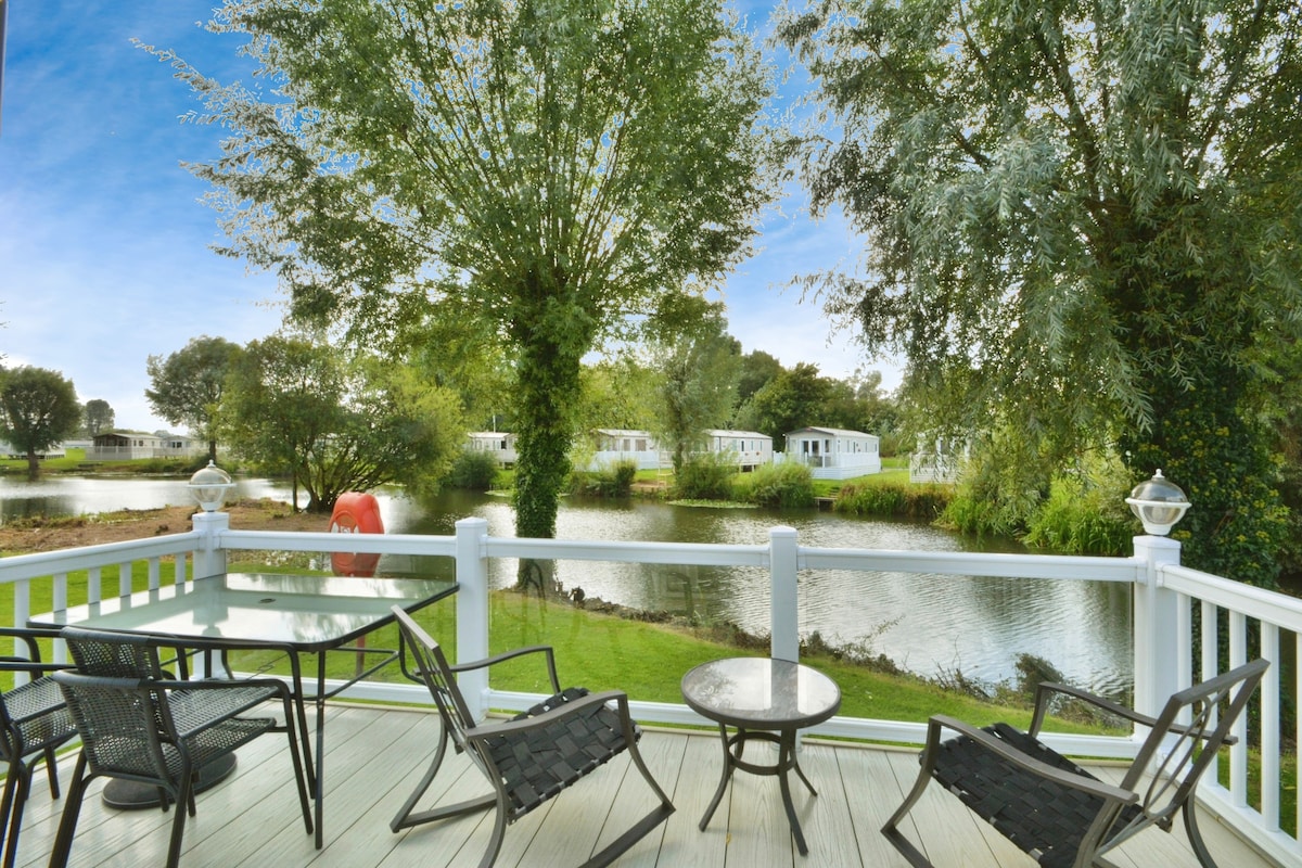 A wooden deck showcases a table and four lounge chairs, providing a serene space for relaxation. Lush greenery surrounds the area, and a tranquil lake reflects the sky, with additional lodges visible in the background. The scene invites moments of quiet contemplation.