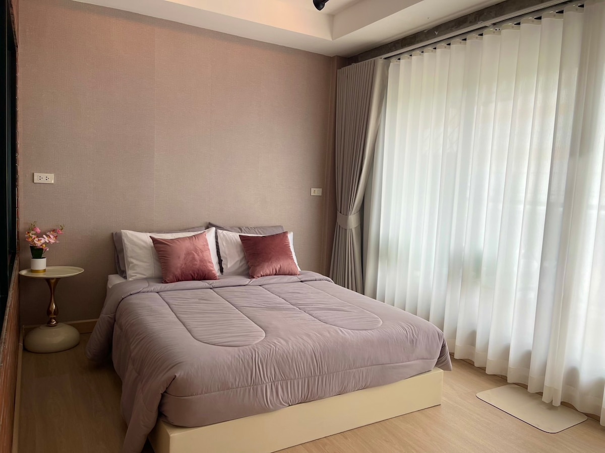 A well-appointed bedroom features a spacious bed with soft, muted bedding and two decorative pillows. Natural light filters through sheer, white curtains, highlighting the room's serene ambiance. A small bedside table with a vase of flowers adds a touch of elegance.