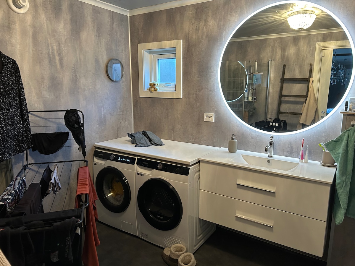 A modern laundry room features a set of front-loading washing machines beside a sleek white countertop. A round mirror is mounted above the countertop, reflecting natural light from a nearby window. A drying rack holds various pieces of clothing, adding functionality to the space.