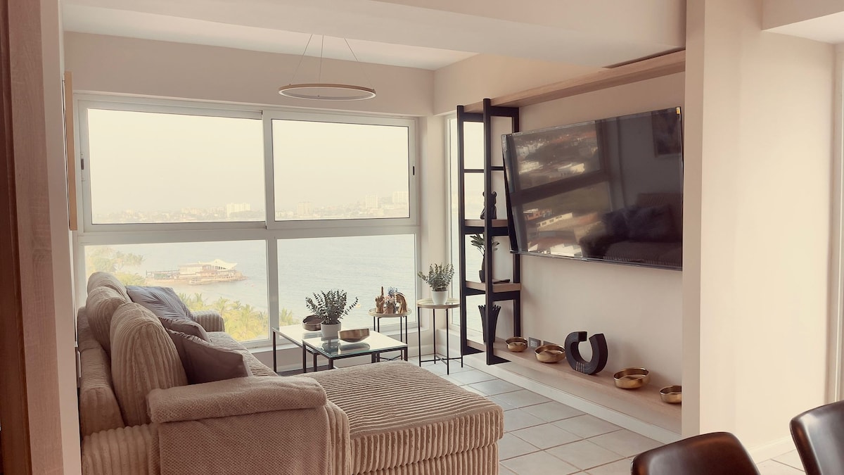 A light-filled living area is presented with large windows offering views of the water. A contemporary, plush sectional sofa is positioned in front of a mounted television. Decorative items are displayed on a sleek coffee table and shelves, enhancing the overall modern aesthetic.