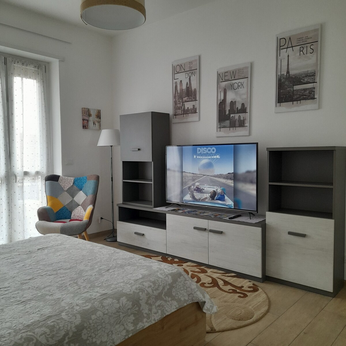 A bright living space features a cozy armchair with a colorful pattern. A television is situated on a stylish media unit, which includes storage shelves. Above, framed city posters enhance the walls, while natural light filters through sheer curtains of a nearby window.