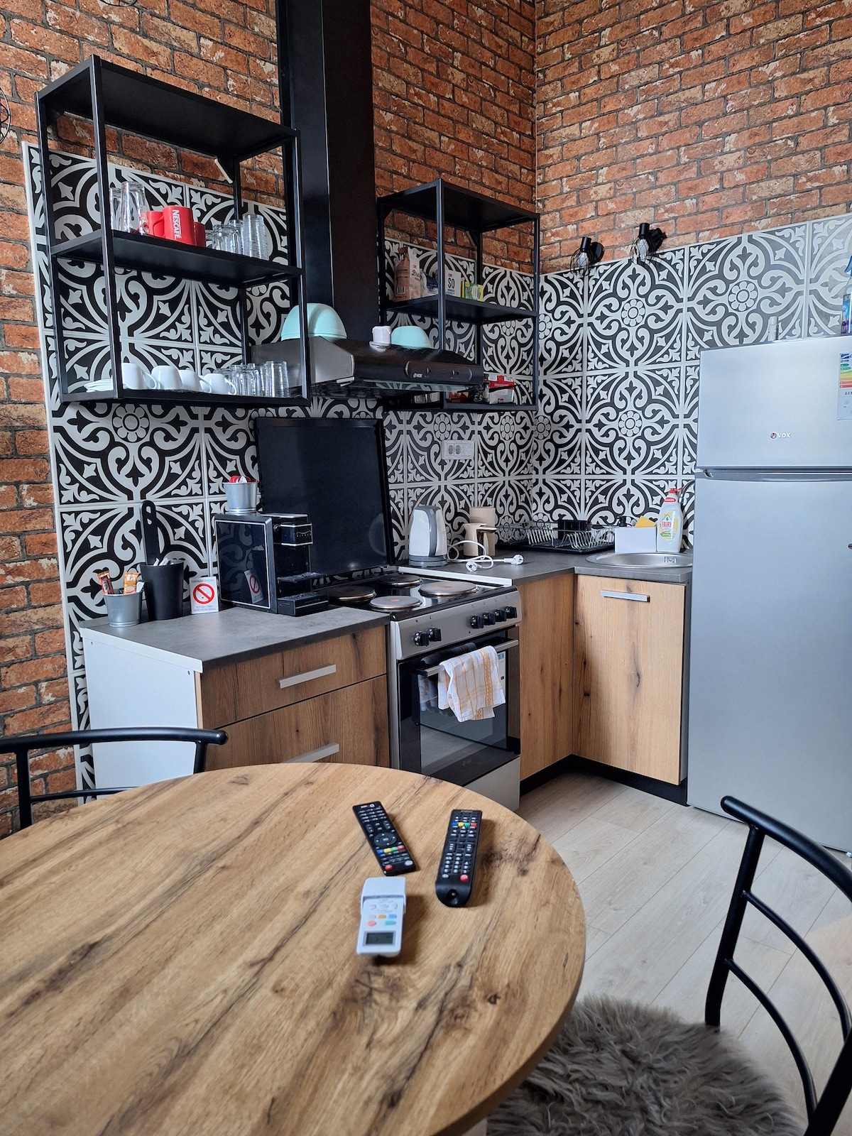 A compact kitchen features a blend of modern appliances and brick decor. Open shelving displays tableware, while a dining table invites gatherings. The kitchen is equipped with a stove, refrigerator, and sink, all set against a patterned tile backsplash.