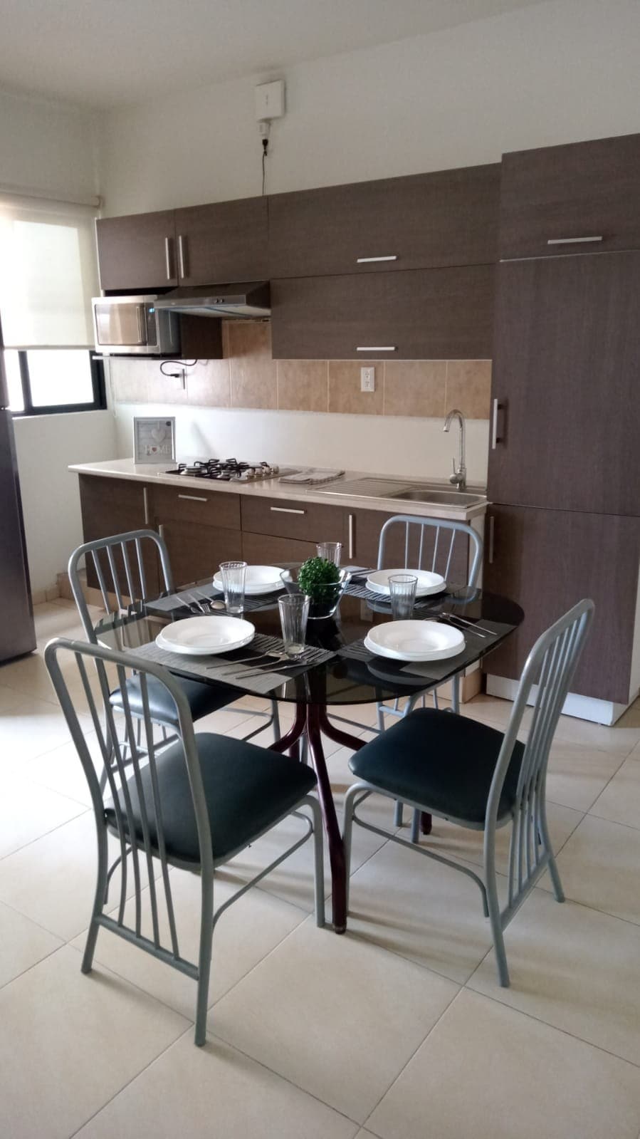 A compact kitchen features modern cabinetry in neutral tones. A dining table set for four is positioned in the center, surrounded by metal chairs. Kitchen appliances, including a stove and microwave, are organized neatly on the countertop, with natural light filtering in through the window.