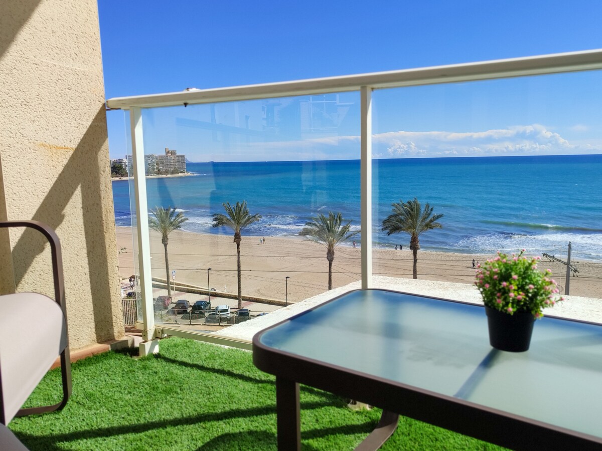 A spacious terrace is framed by a glass railing, offering clear views of the beach and Mediterranean Sea. Palm trees line the shore, and a small potted plant decorates the table. The blue sky enhances the seaside atmosphere, creating a refreshing outdoor space.
