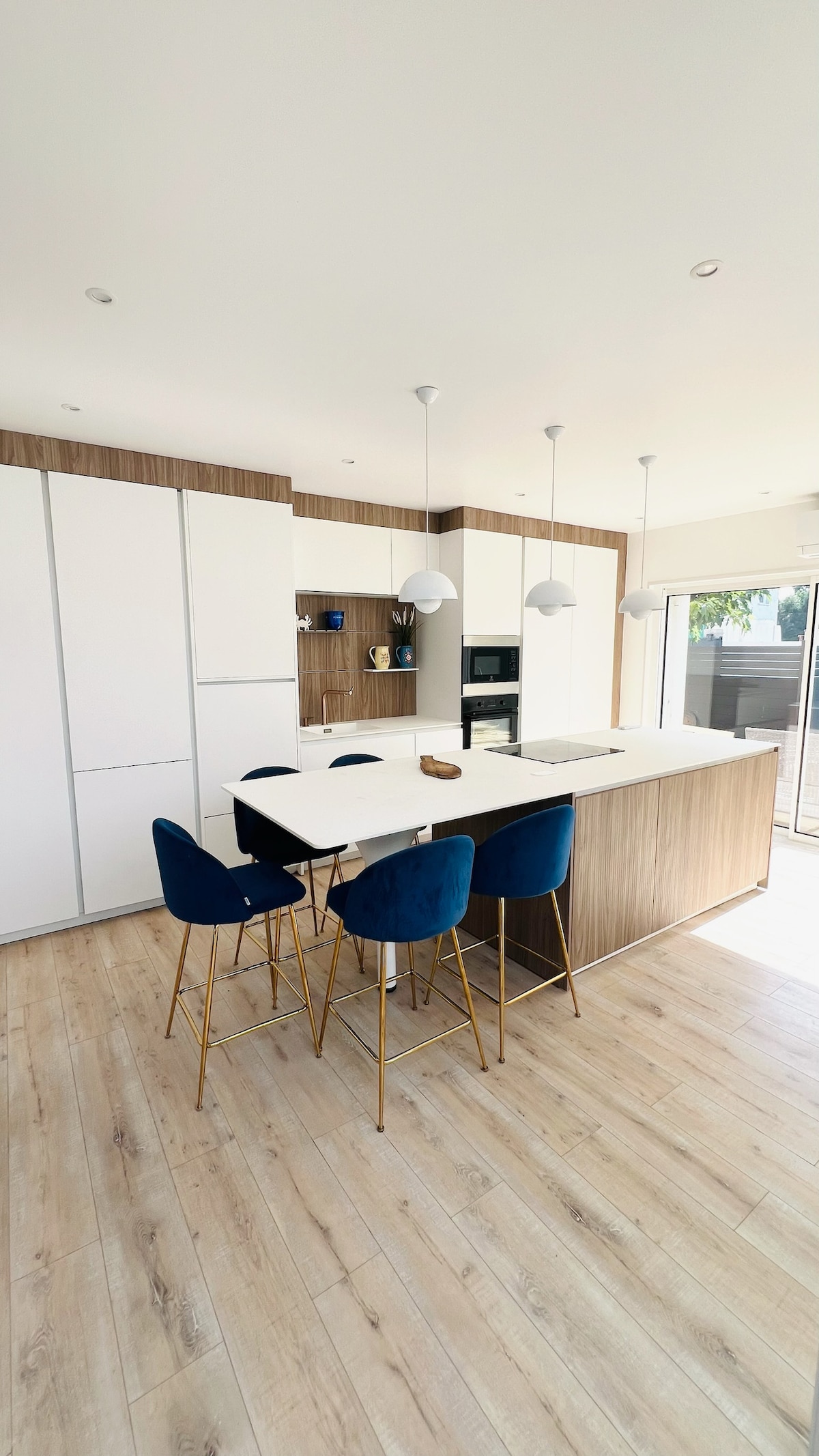 A modern kitchen features a large island with a white countertop and four blue upholstered bar stools. Light wood cabinetry provides storage space, and the room is filled with natural light from nearby windows. White pendant lights hang from the ceiling, illuminating the workspace.