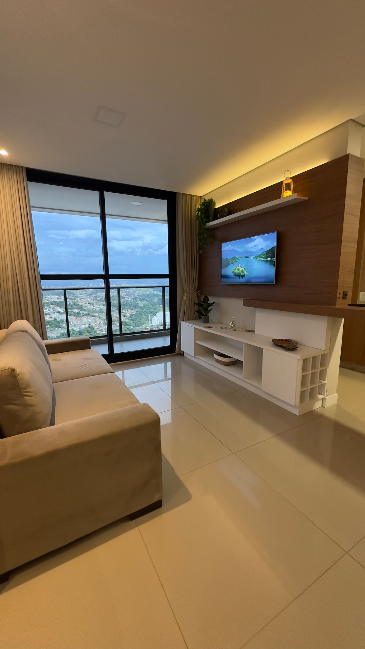 A modern living area features a beige sofa positioned near a large glass window. Natural light floods the space, showcasing scenic views of the surrounding landscape. A television is mounted on a wooden accent wall, complemented by a minimalistic white media console.