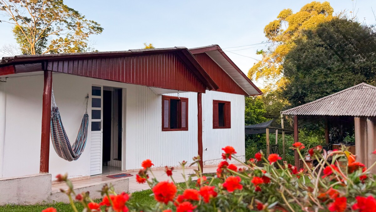 The exterior of the house is shown, featuring a welcoming entrance with a hammock and two windows framed in wood. A covered outdoor area is visible nearby, surrounded by vibrant flowers, highlighting the serene natural surroundings.