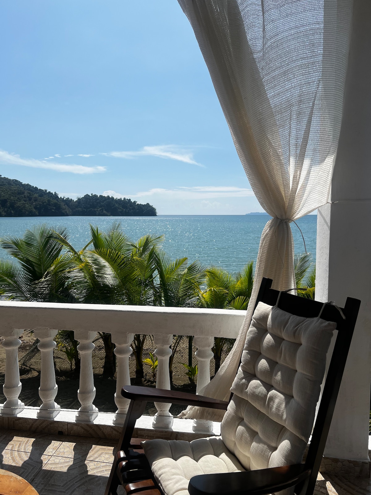 A rocking chair is positioned on a balcony, offering a view of the turquoise waters and lush greenery. Light curtains gently sway in the breeze, enhancing the serene atmosphere created by the tranquil coastal landscape.