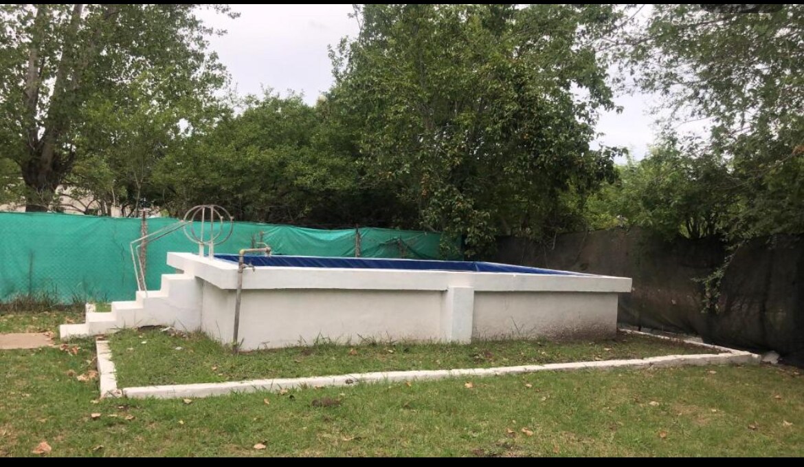 A private swimming pool is surrounded by a well-maintained grassy area. Tall trees provide shade around the fenced pool area, and a set of stairs leads into the pool. A green mesh fence offers privacy while allowing glimpses of the natural surroundings.