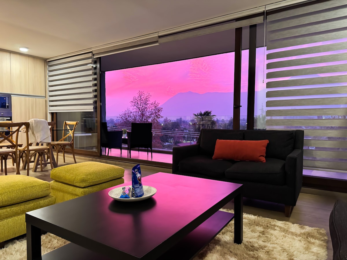 A spacious living area features a modern black sofa and a coffee table at its center. Large glass doors provide an uninterrupted view of the mountains, capturing a colorful sunset. A cozy area rug and a light wooden dining table enhance the inviting atmosphere.