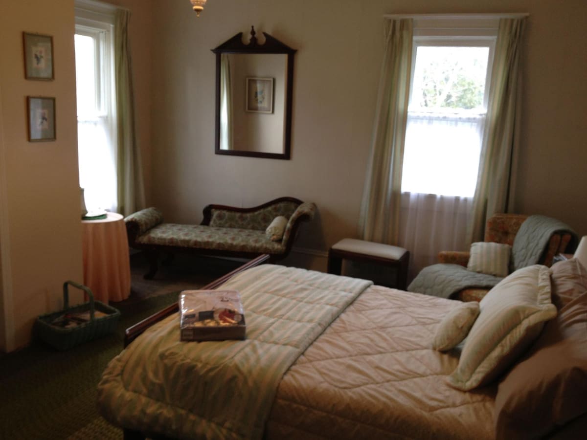 A spacious room features a large bed with soft bedding and several pillows. A vintage settee and armchair are positioned by the windows, allowing natural light to fill the space. Light-colored curtains frame the windows, while framed artwork and a mirror adorn the walls.