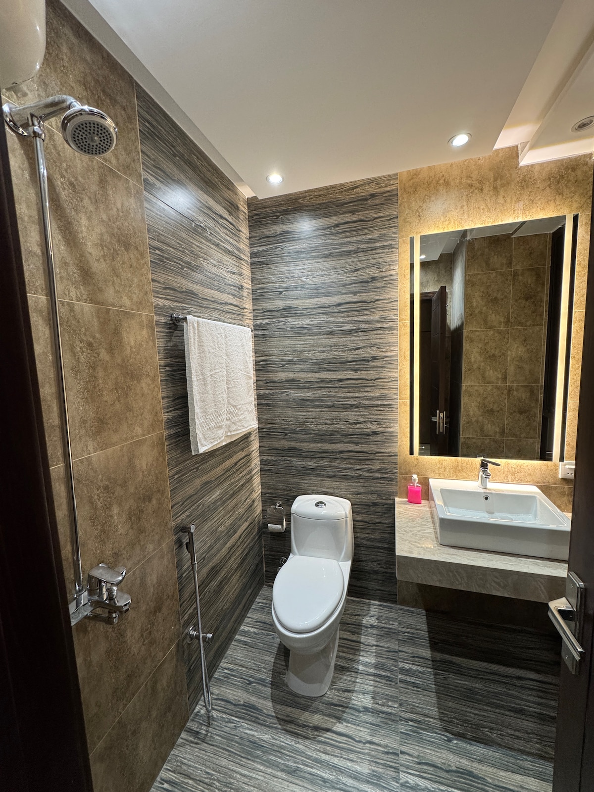The bathroom features a modern design with textured wall tiles in neutral tones. A sleek white toilet and a minimalistic vanity with a sink are visible. Soft lighting accentuates the space, complemented by a large mirror and a towel hung on the wall.