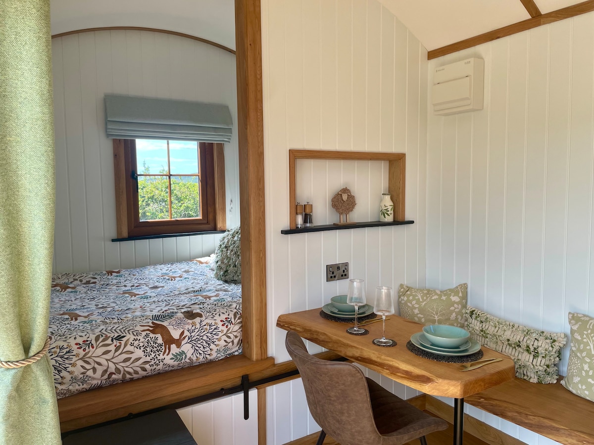 An inviting interior showcases a spacious bed adorned with a patterned duvet, framed by a light window. A wooden table is set for two, featuring plates and glasses, while a small shelf displays decorative items. Soft green curtains add a touch of warmth.