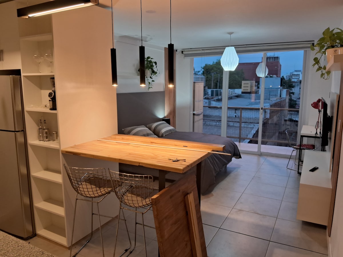 A modern studio apartment features a spacious sleeping area with a large bed, complemented by a work desk and an open kitchen setup. The space is illuminated by pendant lights, with large windows offering city views and allowing natural light to fill the room.