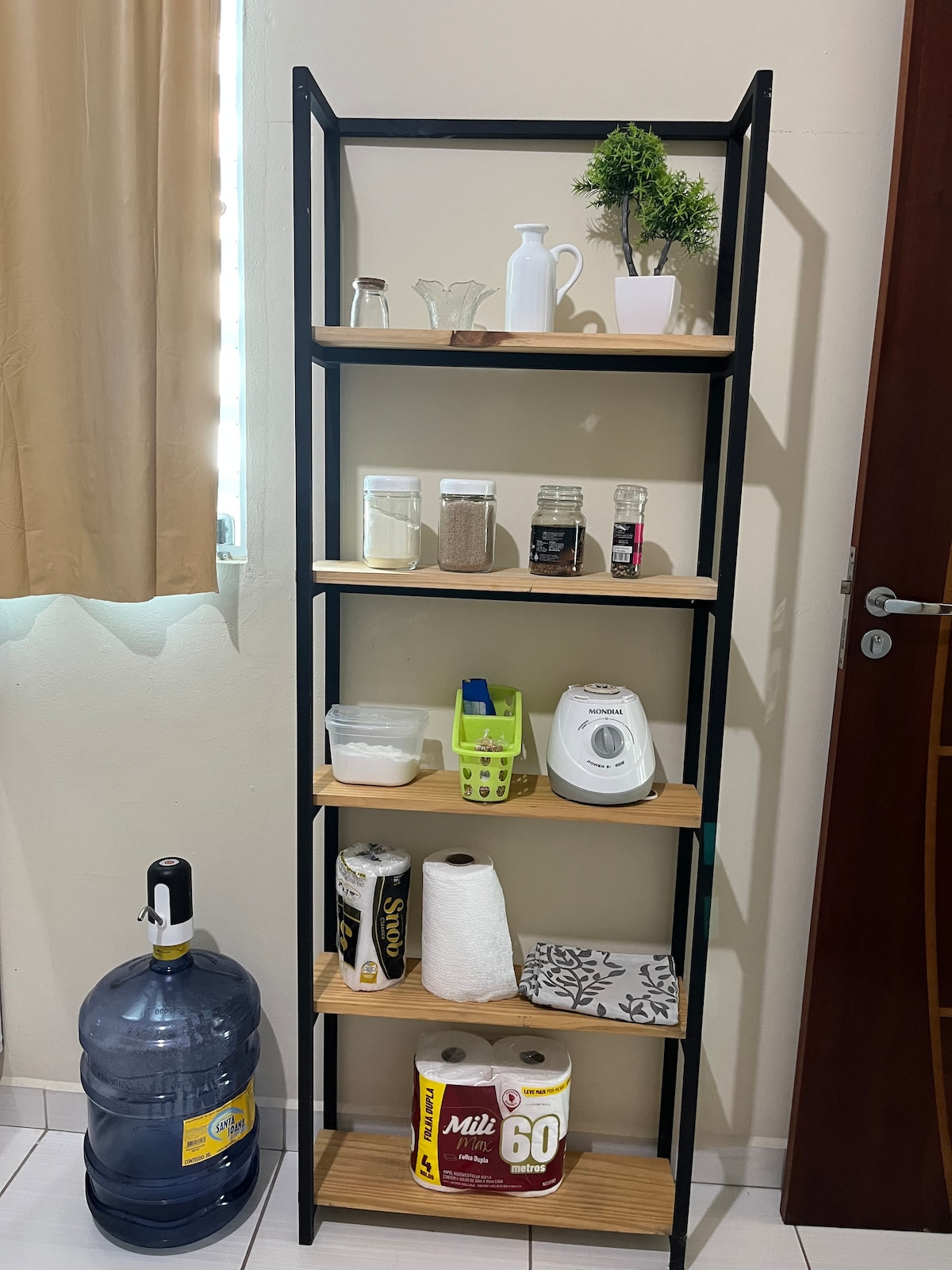 A multi-tiered shelving unit is displayed against a light wall, featuring various items such as glass containers, a small plant, and kitchen essentials. A large water dispenser rests on the floor beside the shelf. Soft natural light enters through a nearby window with light-colored curtains.