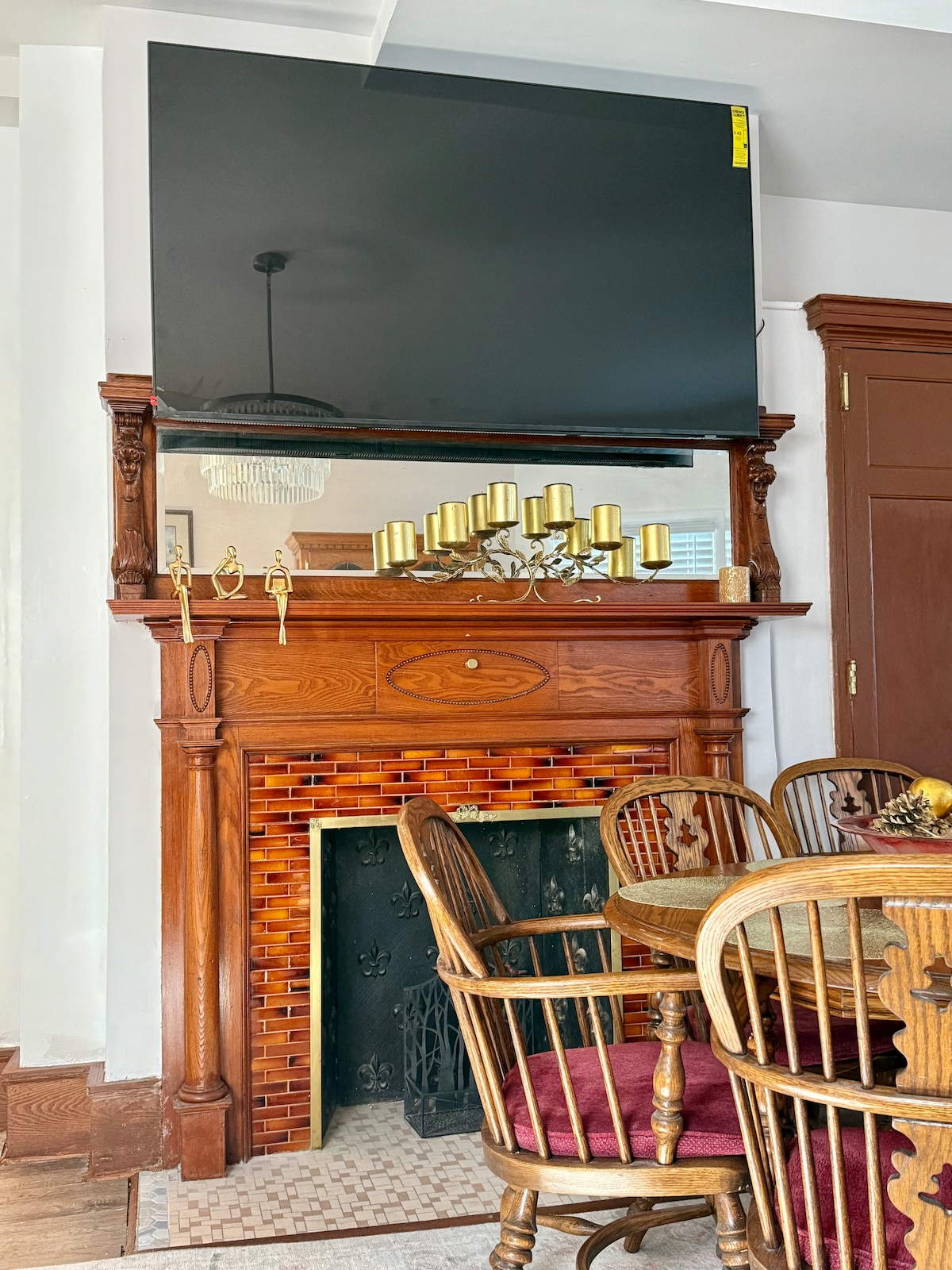 An elegant dining area displays a wooden dining table surrounded by matching chairs. Above the ornate fireplace, a large flat-screen television is mounted. The fireplace features decorative tiles, and a chandelier adds a touch of sophistication to the space.