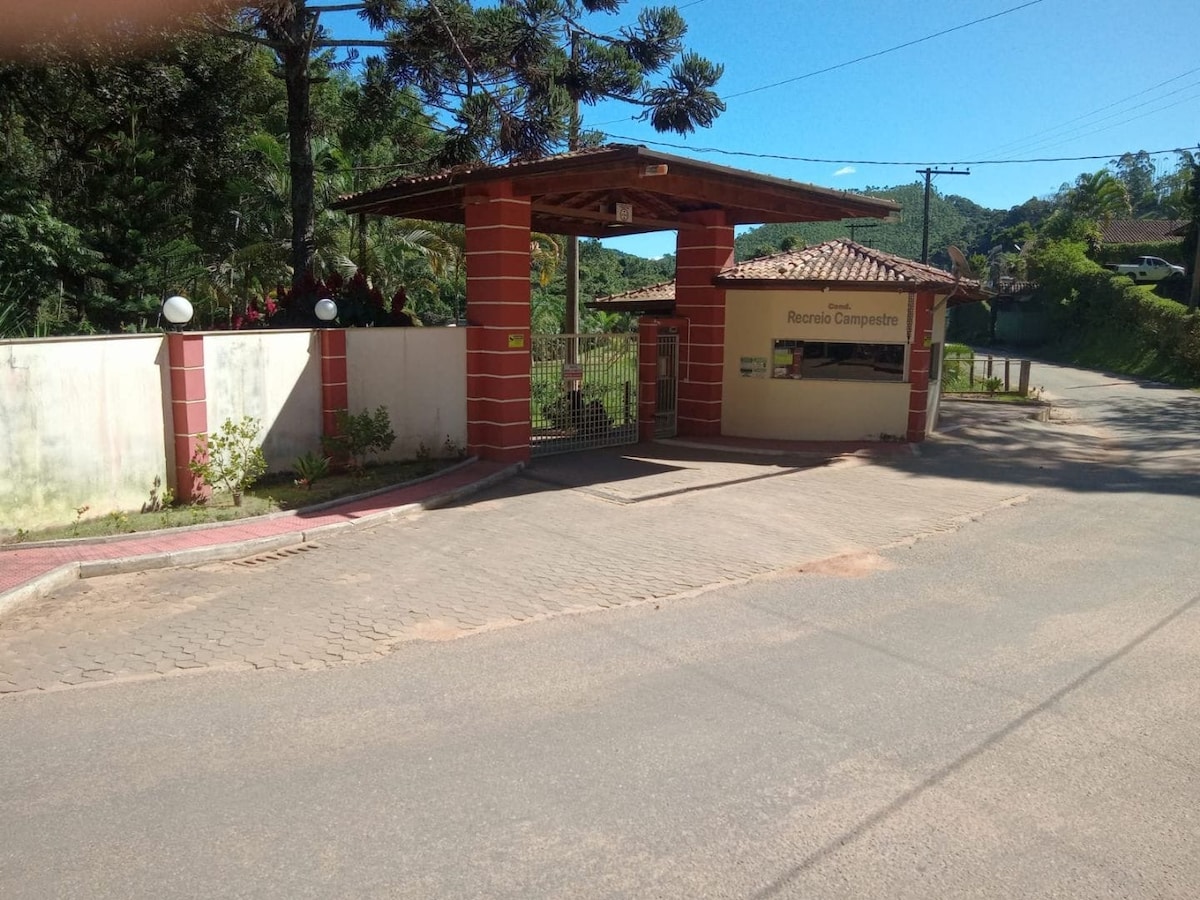 The entrance to a private condominium is shown, featuring a gated guardhouse and surrounding lush greenery. The roadway leads into the community, with a well-maintained access path and ample landscaping evident around the entrance.