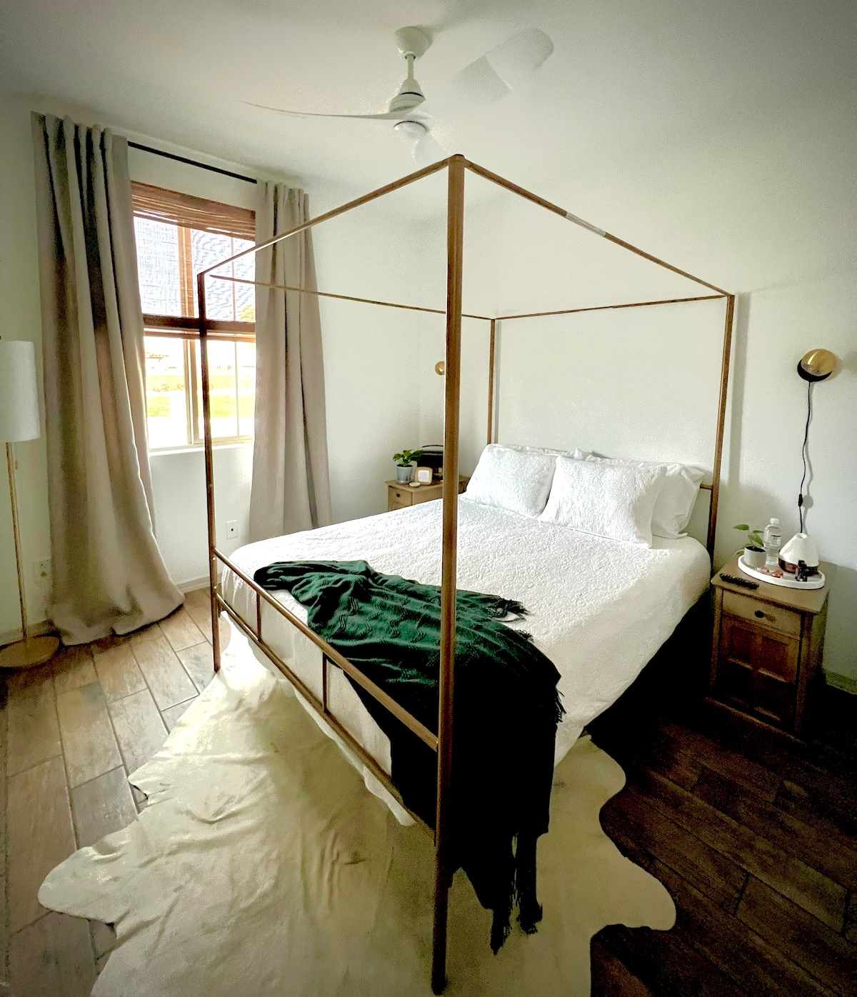 A calm bedroom features a queen size canopy bed draped with a dark green throw blanket. Natural light enters through a large window adorned with light curtains. A small nightstand with a lamp and a plant adds subtle warmth to the space.