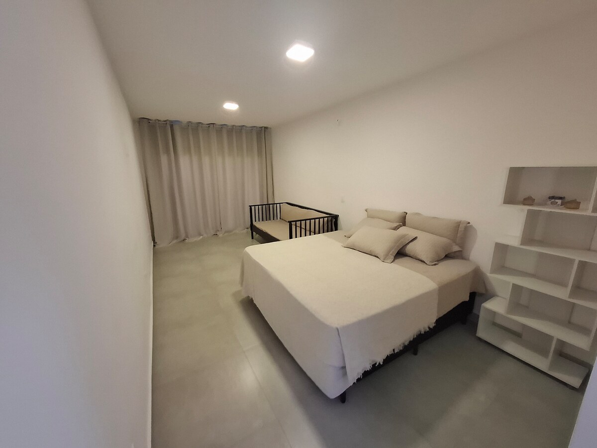 A spacious bedroom is equipped with a large bed covered in neutral linens and accompanied by decorative pillows. A minimalist crib is positioned nearby, and natural light filters through the sheer curtain. A simple shelf is visible against the wall, contributing to the room's clean aesthetic.