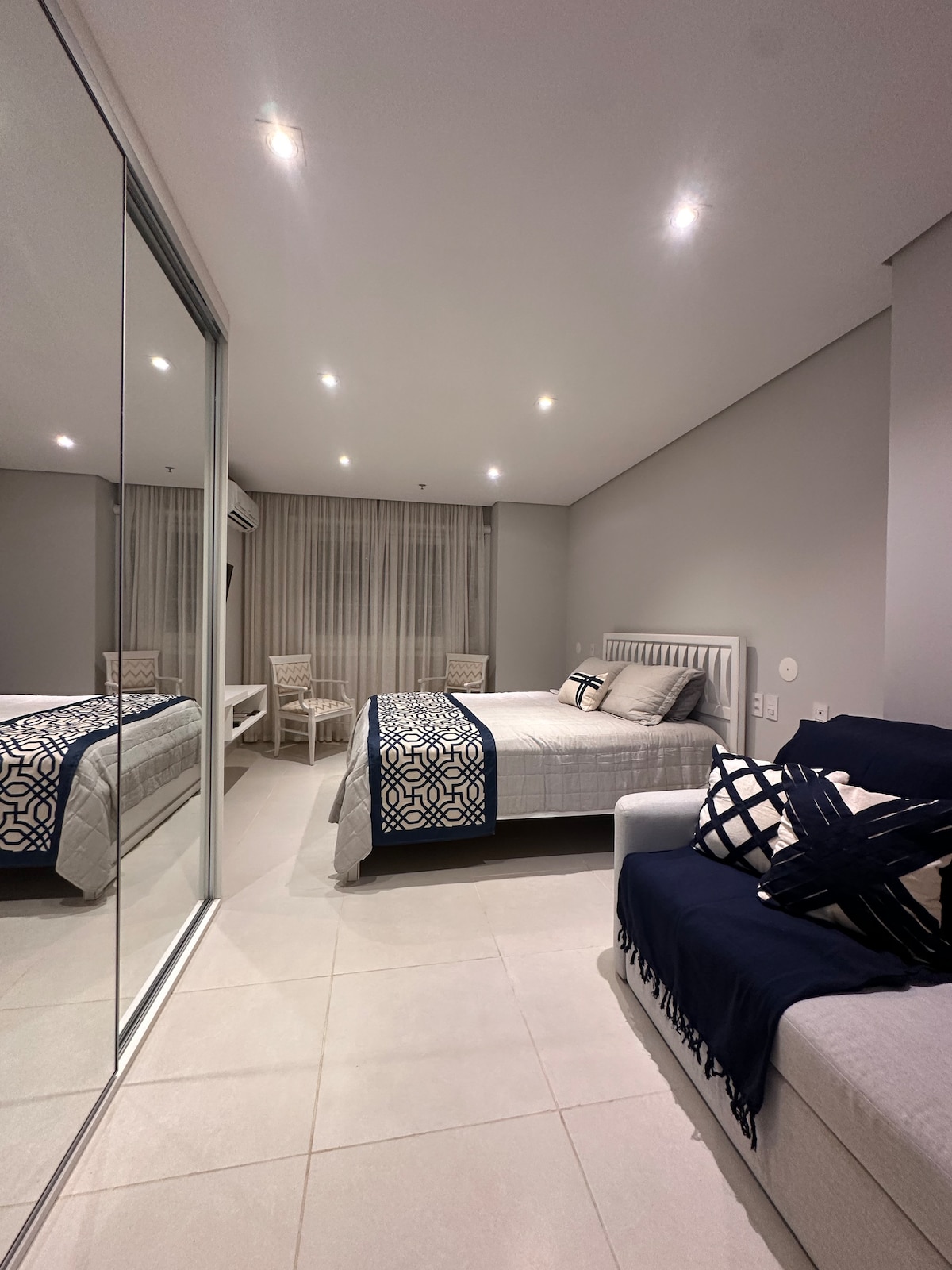 A well-appointed sleeping area features a spacious bed with decorative pillows. A white sofa with patterned cushions is positioned nearby. The room boasts large mirrored wardrobes and soft lighting, creating an airy ambiance. Floor-to-ceiling windows are draped with light sheer curtains.
