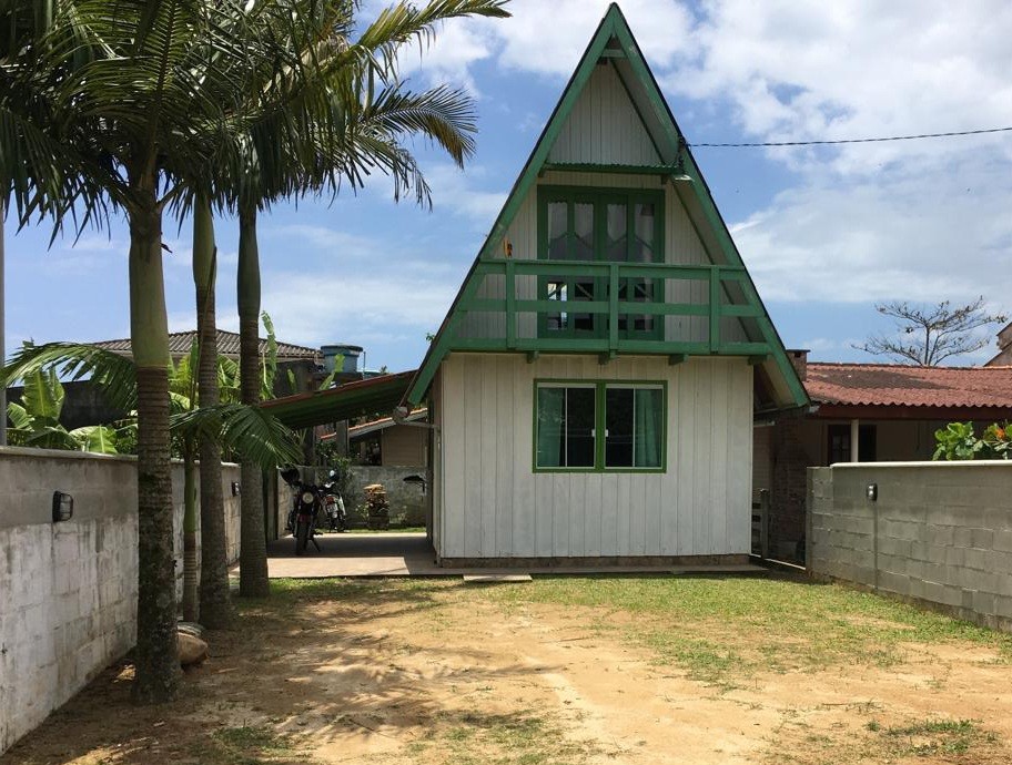 A charming A-frame chalet is set against a clear sky, featuring green accents on the balcony and windows. A grassy area surrounds the structure, providing a sense of openness. Palm trees are positioned on one side, adding to the tranquil atmosphere of the property.