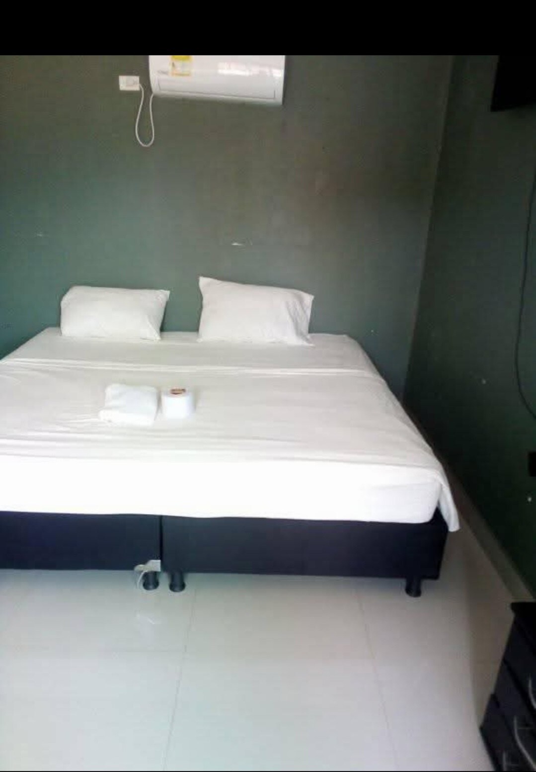 A cozy sleeping area features a spacious bed dressed in crisp white linens, with two pillows placed neatly at the head. A white towel is arranged on the bed, while the soothing green walls provide a calming backdrop.