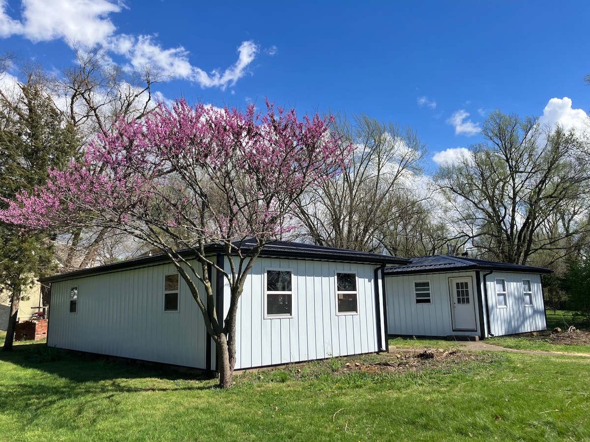 A single-story structure with a light-colored exterior is surrounded by lush green grass. A flowering tree with pink blossoms is positioned in the foreground, complementing the serene setting. The clear blue sky and scattered clouds add to the peaceful atmosphere of the location.