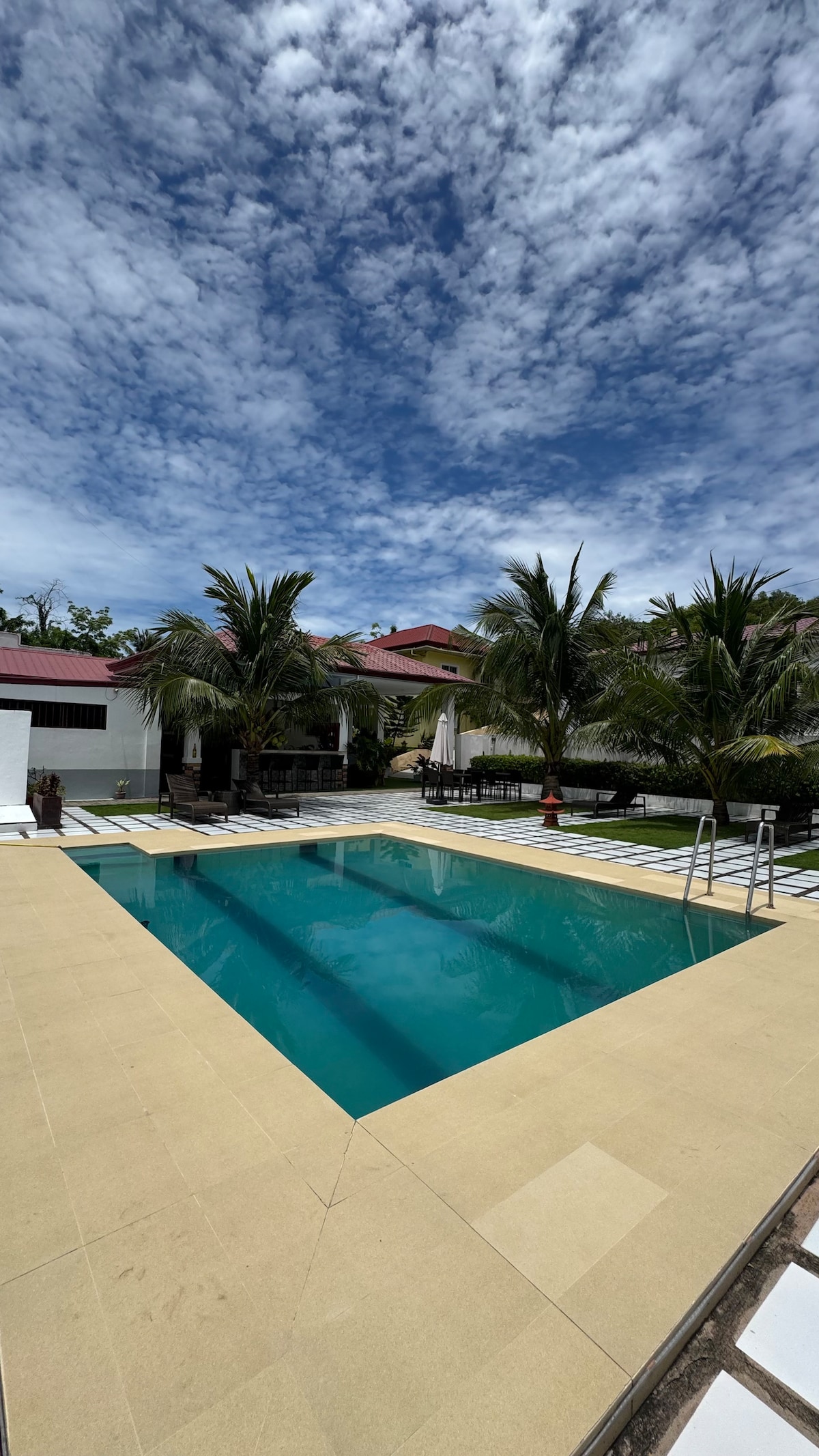 A tranquil swimming pool is framed by a spacious patio and surrounded by palm trees. The clear water reflects the blue sky, while the pool's edge features a light-colored tile surface. Nearby structures can be seen providing shade and comfort.