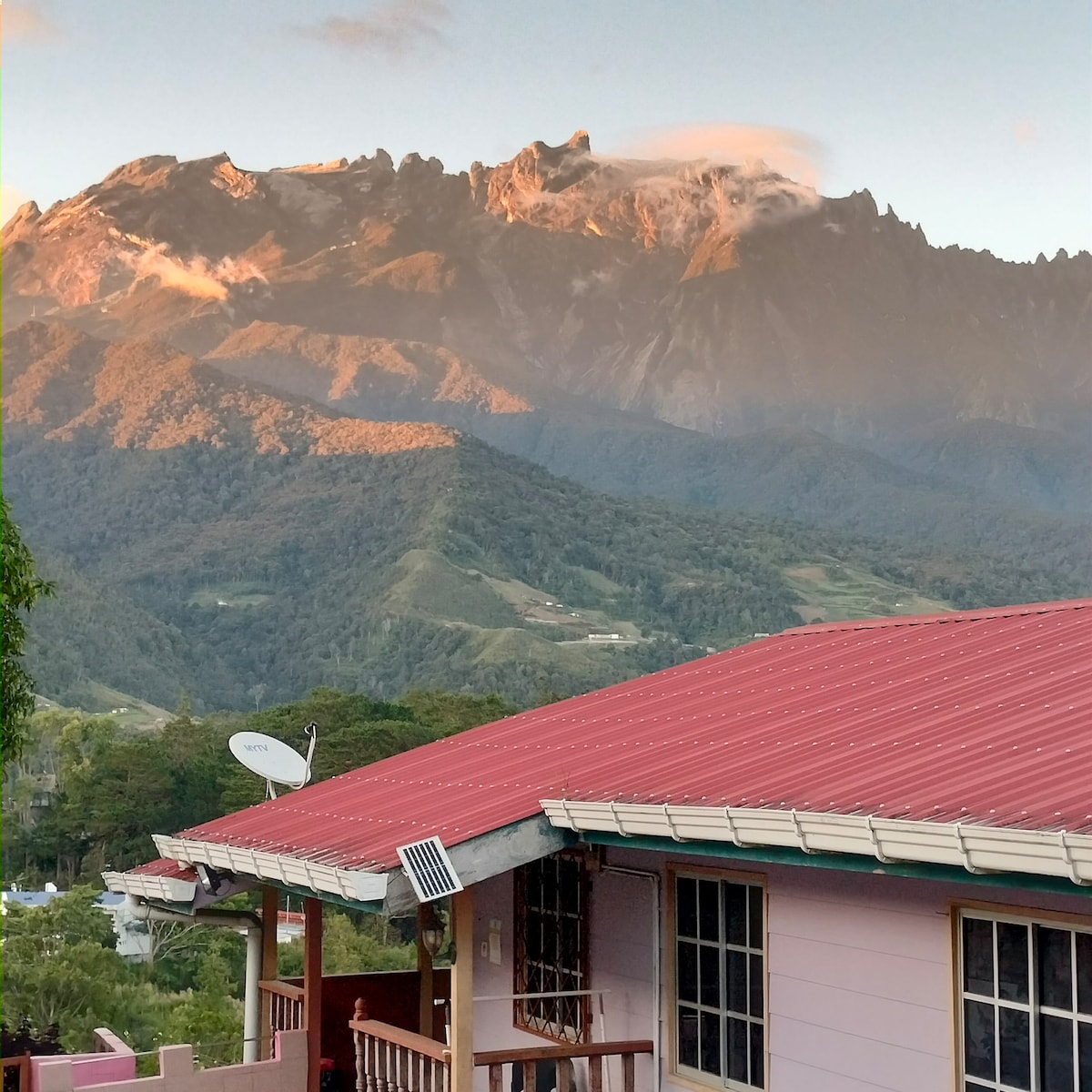 A view of a house with a red roof is showcased against a backdrop of majestic mountains. The peaks appear to be touched by sunlight, while a mix of trees covers the lower slopes. A satellite dish is mounted on the side of the house.