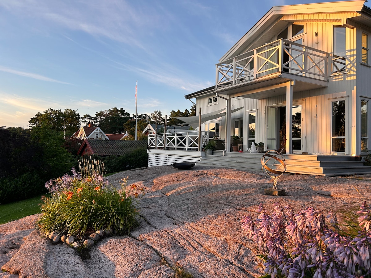 The image depicts a modern villa with a spacious deck, set on a rocky terrain. Large windows invite natural light, and colorful flowers add a touch of vibrancy to the landscape. The surrounding area features lush greenery and glimpses of neighboring structures in a tranquil setting.