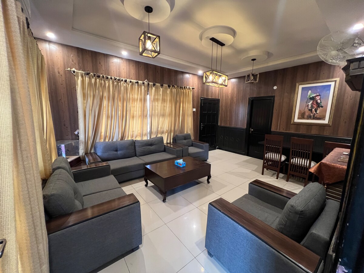 A spacious living area features grey upholstered couches arranged around a central wooden table. Natural light filters through long, light-colored curtains, accentuating the wooden wall paneling. A dining area with wooden chairs is visible in the background alongside warm pendant lighting.