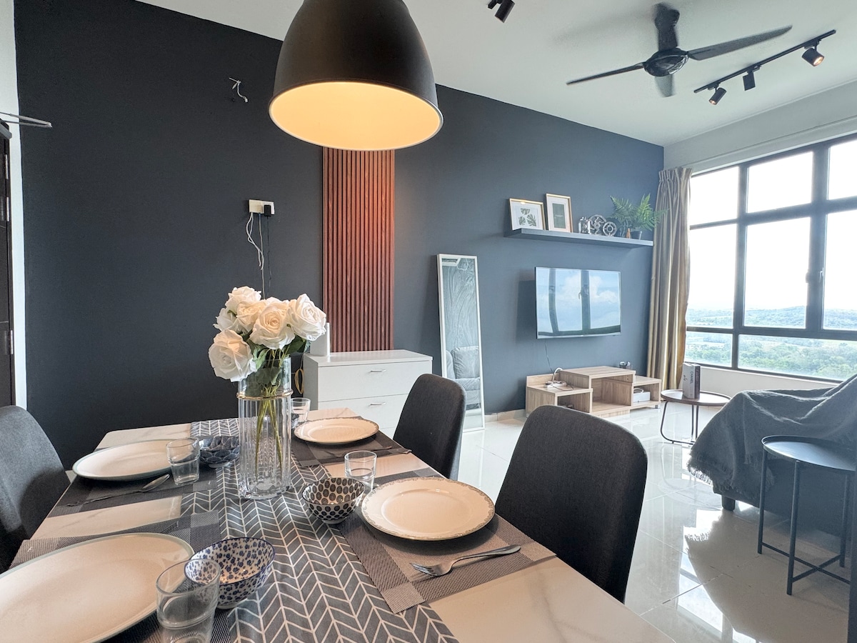 The dining area features a modern table set for four, complemented by decorative plates and glassware. A pendant light illuminates the space, while a stylish lounge area with a television and a bed in the background offers a glimpse of the expansive view through large windows.