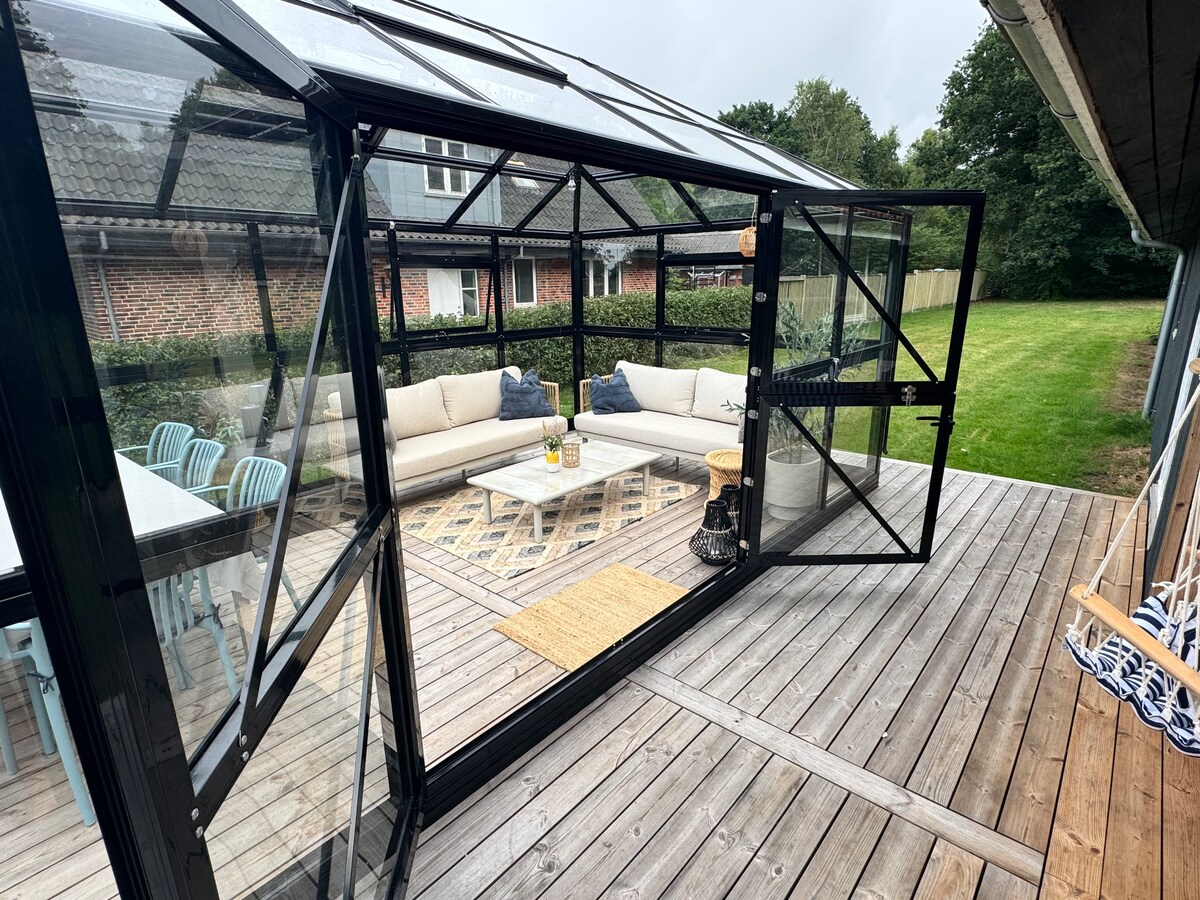 A spacious glass conservatory is shown, featuring a lounge area with cozy seating and a central coffee table. The structure is surrounded by sliding doors, providing easy access to a wooden deck. A grassy area is visible in the background, enhancing the outdoor experience.