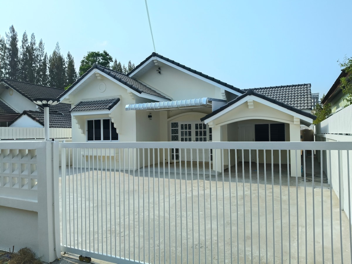 A newly renovated house features a modern exterior with a clean white facade. A spacious driveway is visible, along with a gated entrance. Surrounding greenery adds a touch of nature to the front of the property.