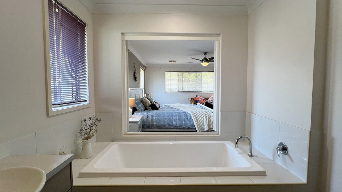 The image displays a spacious bathroom that includes a large white bathtub. A window is framed by wooden blinds, offering a view of a well-lit bedroom featuring a kingsize bed. The modern simplicity of the space is enhanced by neutral-toned walls.