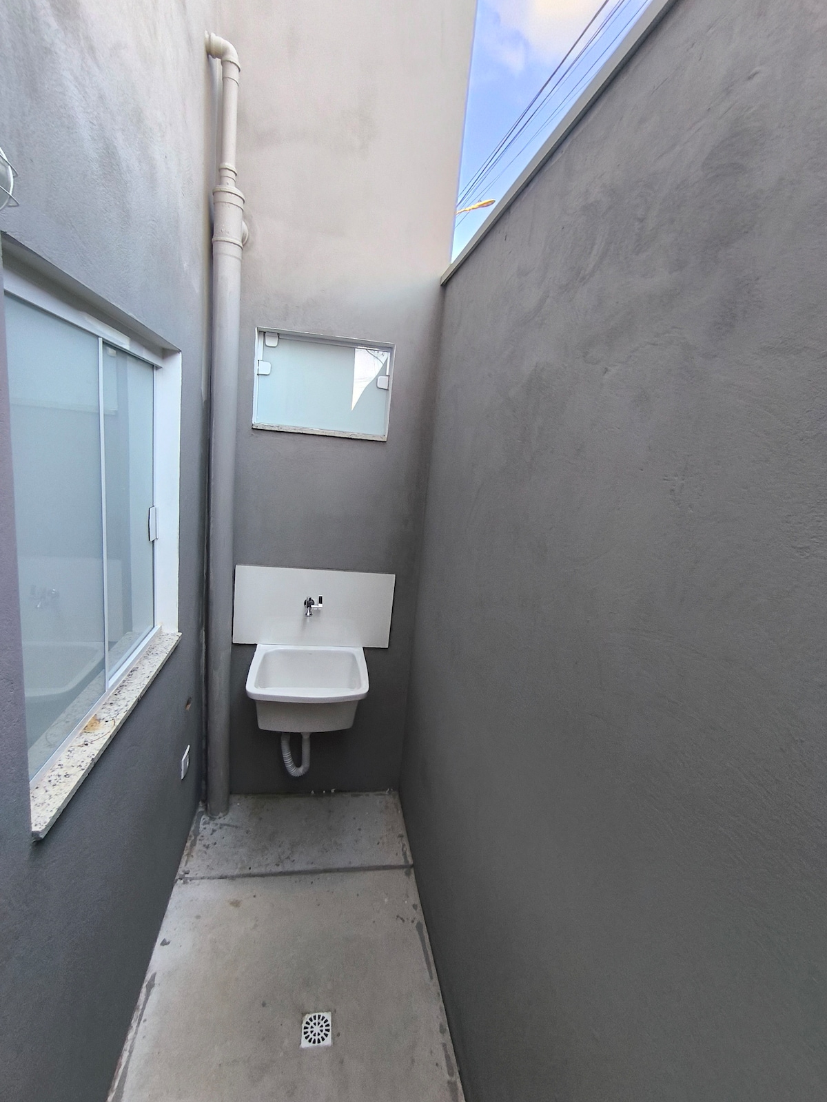 A narrow outdoor space is shown, featuring light gray walls and a simple floor. A white sink is mounted to the wall, accompanied by a small mirror above it. Natural light enters through a window on the side, brightening the area.