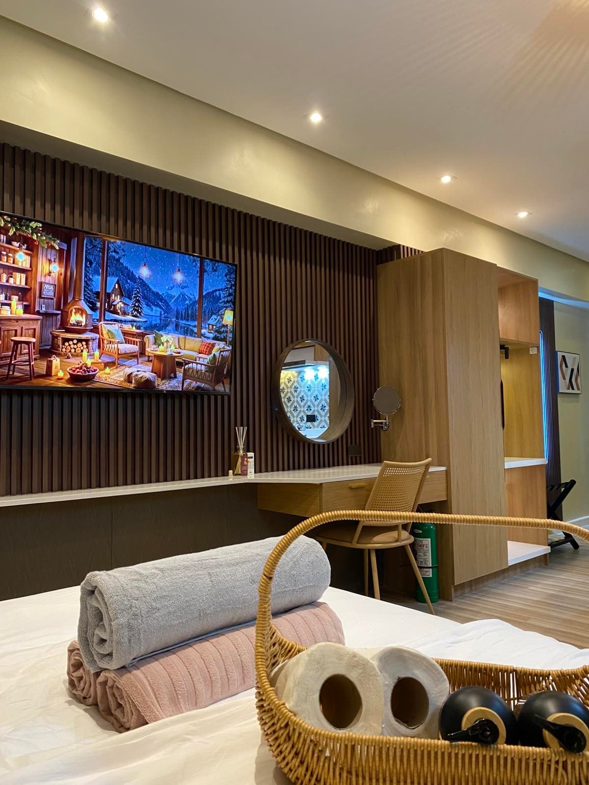 A studio room is depicted, featuring a wall-mounted television above a wooden desk area. A round mirror, a wicker basket with toiletries, and neatly rolled towels on the spacious bed enhance the inviting ambiance. Soft lighting highlights the textured wall and modern decor.