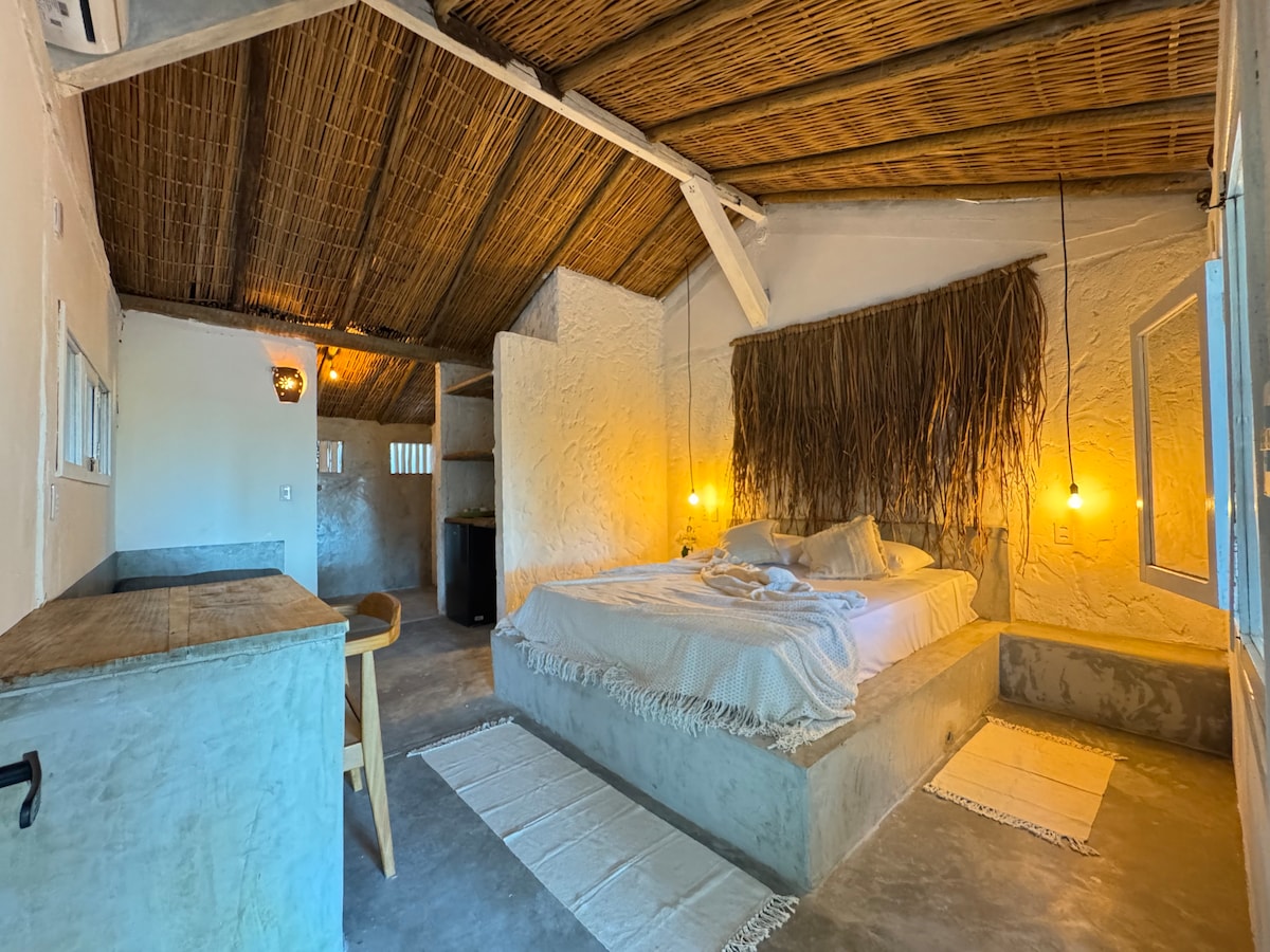 A spacious bedroom features a queen-sized bed with soft linens, illuminated by warm pendant lighting. Textured natural walls complement a thatched ceiling. A rustic wooden table and chair are positioned near a window, offering a serene atmosphere. Soft woven rugs add comfort to the concrete flooring.