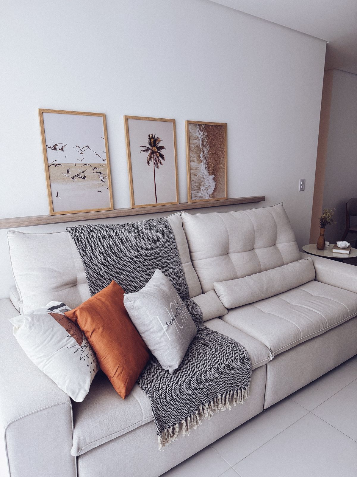 A light-colored sofa is complemented by decorative cushions, including a textured gray one and an orange accent pillow. Above the sofa, three framed artworks depict beach scenes, adding a serene touch to the room. A small side table is visible, enhancing the cozy atmosphere.