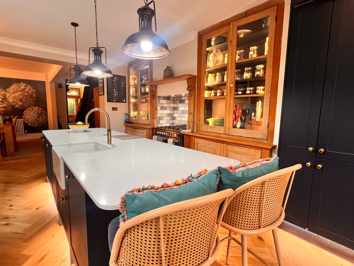 An open-plan kitchen showcases a large island with a white countertop and decorative seating. Wooden cabinets with glass display doors contain various items. Soft lighting is provided by pendant lamps above the island, illuminating the inviting space.