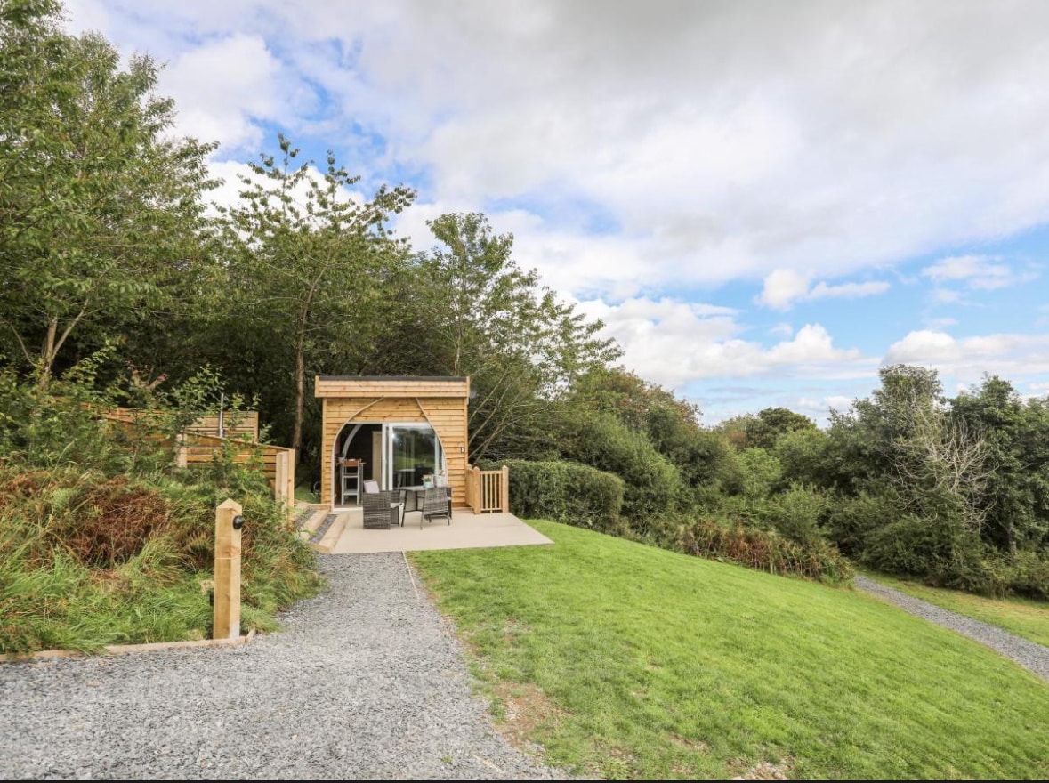 A wooden pod structure is shown set against a natural backdrop of trees and greenery. A small outdoor seating area with table and chairs is visible on a paved patio, surrounded by soft grass and gravel paths leading up to the entrance.