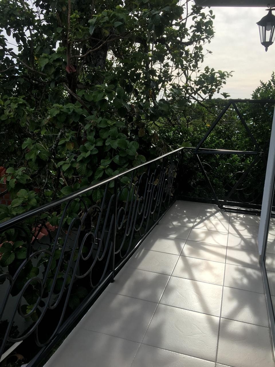 A balcony is framed by lush greenery, offering a sense of privacy and tranquility. A decorative wrought-iron railing outlines the space, and the tiled flooring enhances the open feeling. Natural light softly illuminates the area, creating a serene retreat.