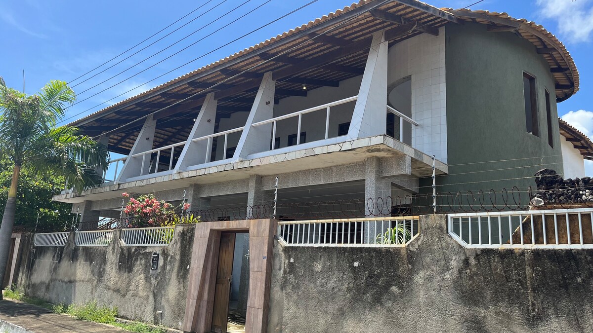 The exterior of the beach house showcases a spacious two-story design with a large covered balcony, framed by light-colored walls and a distinctive green facade. Lush foliage and flowering plants enhance the inviting entry, while the structure is complemented by a tiled roof.