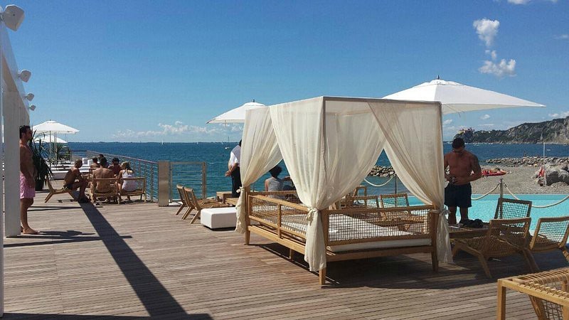 A beachfront area features wooden decking with several lounge chairs and a shaded cabana. Umbrellas are positioned throughout, providing shade. The sea is visible in the background, reflecting calm blue waters under a clear sky, with people leisurely enjoying the space.