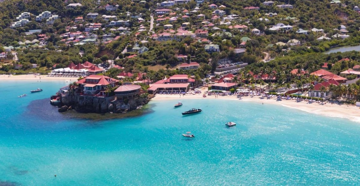 Aerial view of a serene beach area with clear turquoise water, bordered by soft sand. Beachfront establishments with red rooftops blend into the lush hillside. Boats are anchored in the water, creating a calm and inviting coastal scene.