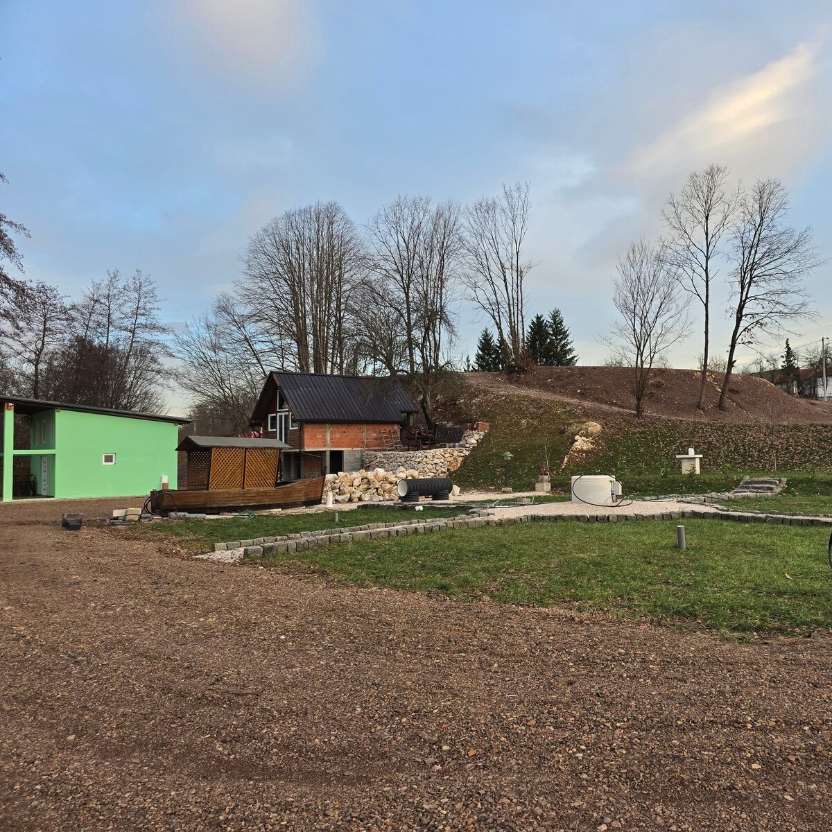 A serene outdoor setting features a rustic cabin with a dark roof, complemented by a nearby green structure. A grassy area surrounds the cabin, with a gravel pathway visible. Trees and a hillside in the background enhance the natural landscape.