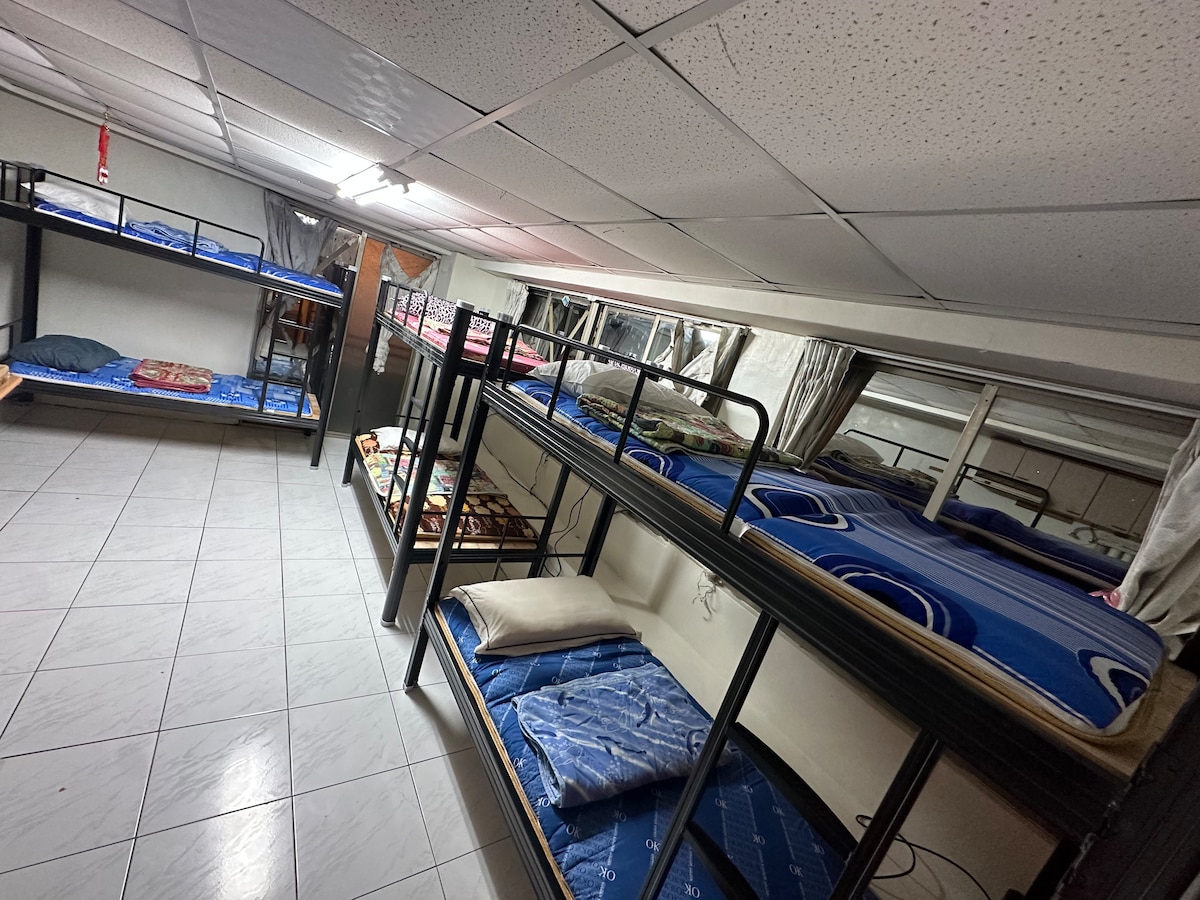A shared sleeping area displays multiple bunk beds aligned against the walls, featuring various bedding patterns. Some mattresses are neatly arranged, while others show partial blankets. A bright ceiling light illuminates the room, highlighting the tiled floor and the windows covered with light curtains.